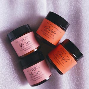 May include: Four jars of body butter with black lids and labels. Two jars have pink labels with "Warm Spice" text, and two have orange labels with "Citrus Wood" text. The jars are arranged on a white, textured surface.