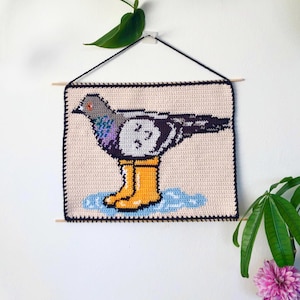 Op de afbeelding: Gehaakte wanddecoratie met een duif in gele regenlaarzen, staand in een plas. De vogel is grijs, zwart en wit, met paarse accenten. De achtergrond is beige en de rand is zwart. Het kunstwerk is opgehangen met een zwart koord en houten stokken.