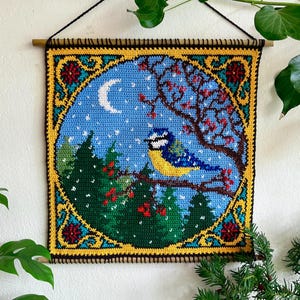 May include: A square, crocheted wall hanging featuring a blue bird perched on a branch in a winter scene. The artwork includes a crescent moon, snow, and a tree with red berries, framed by a yellow and black border. The piece is suspended from a wooden dowel.