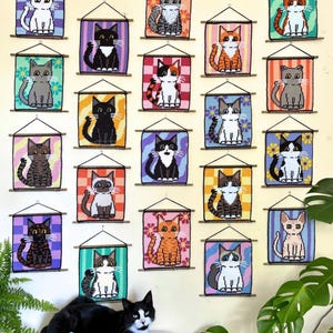 Puede incluir: Una colección de veintiún tapices rectangulares con diversas ilustraciones de gatos en variados colores y patrones, cada uno suspendido de un listón de madera con un cordón negro, dispuestos en una cuadrícula sobre una pared de color claro.