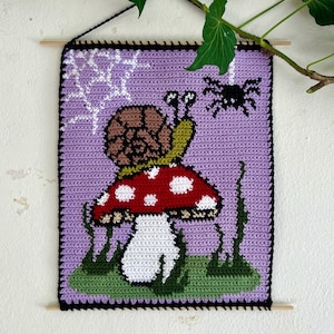 Könnte beinhalten: Gehäkeltes Wandbild mit einer Schnecke auf einem rot-weiß gepunkteten Pilz, einer schwarzen Spinne und einem Spinnennetz auf lila Hintergrund. Das Kunstwerk ist mit einem schwarzen Rand eingefasst und hat eine Holzstange zum Aufhängen.