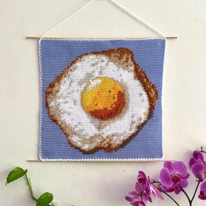 May include: A crocheted wall hanging featuring a fried egg design. The egg has a yellow yolk and a white egg white with a brown, crispy edge, set against a light blue background. The artwork is suspended by a white string and wooden dowels.