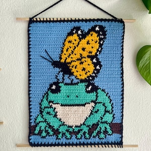 May include: Crocheted wall hanging featuring a frog with a butterfly on its head. The frog is green and teal, with a cream-colored belly, perched on a brown branch. The butterfly is yellow and black, set against a light blue background.