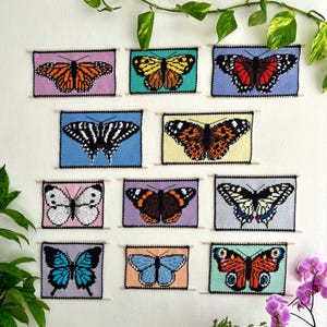 May include: Eleven framed butterfly needlepoint artworks are displayed on a white wall. Each piece showcases a unique butterfly design in colours such as orange, blue, and black. The wall is decorated with green plants.