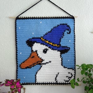Könnte beinhalten: Gehäkeltes Wandbild mit einer weißen Ente, die einen blauen Hexenhut mit gelbem Band trägt. Die Ente hat einen orangefarbenen Schnabel und ist vor einem hellblauen Hintergrund mit weißen Sternakzenten platziert. Das Kunstwerk ist mit einem schwarzen Rand eingefasst.