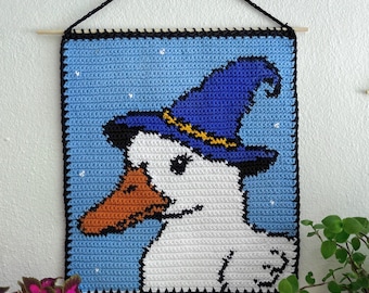May include: A crocheted wall hanging depicting a white duck wearing a blue witch's hat with a yellow band. The duck has an orange beak, set against a light blue background with white star accents. The artwork is framed with a black border and hangs from a wooden dowel.