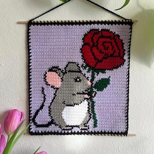 Könnte beinhalten: Gehäkeltes Wandbild mit einer grauen Maus, die eine rote Rose vor einem lavendelfarbenen Hintergrund hält. Das Kunstwerk ist mit einem schwarzen Rand und Holzstäben zum Aufhängen versehen. Die Maus hat rosa Ohren und einen weißen Bauch.