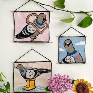 Puede incluir: Tres tapices de pared hechos a mano con ganchillo con diseños de palomas. Cada obra de arte cuadrada tiene un borde negro y está suspendida de una clavija de madera. Las palomas se representan en varias poses, con diferentes fondos de colores.