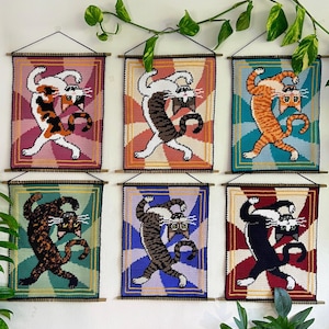 May include: Six colorful cat tapestries. Each tapestry features a cat in a dynamic pose against a geometric background. The cats are in various colors, including calico, tabby, and orange. The tapestries are framed with a wooden rod and black string.