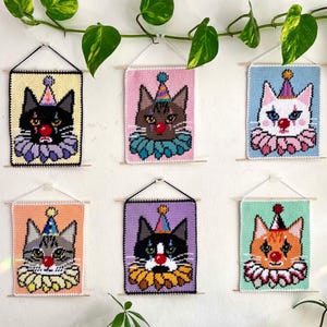 May include: Six colorful, handmade cat portraits hang on a wall. Each cat is depicted as a clown, wearing a party hat and ruffled collar. The portraits are made with yarn and mounted on wooden dowels.