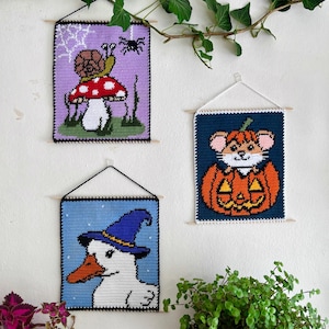 Può includere: Tre arazzi da parete all'uncinetto con disegni stravaganti. Uno presenta una lumaca su un fungo, un altro un topo in una zucca e il terzo un'anatra con un cappello da strega. Ognuno è incorniciato e appeso con un tassello di legno.