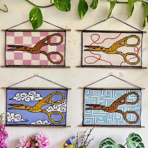 May include: Four needlepoint wall hangings featuring gold scissors. Each piece has a different background: pink and white checkerboard, white with red thread, blue with clouds, and teal geometric pattern. The scissors are the focal point.