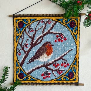 Könnte beinhalten: Gehäkeltes Wandbild mit einem Rotkehlchen auf einem schneebedeckten Ast mit roten Beeren. Das quadratische Design hat einen blauen Hintergrund mit weißen Schneeflocken, umrahmt von einem gelb-blauen Rand. Aufgehängt mit einer Holzstange.