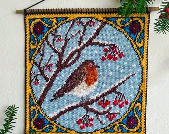 Op de afbeelding: Gehaakte wandhanger met een roodborstje op een besneeuwde tak met rode bessen. Het vierkante ontwerp heeft een blauwe achtergrond met witte sneeuwvlokken, omlijst door een gele en blauwe rand. Opgehangen met een houten roede.