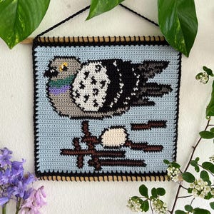 May include: Crocheted wall hanging featuring a pigeon design. The bird is depicted in shades of gray, black, and white, set against a light blue background. The artwork is framed with a black border and a wooden dowel for hanging.