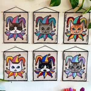 May include: Six handmade cat wall hangings. Each features a cat wearing a colourful jester hat and collar. The word "JOKER" is printed vertically on the sides. The cats have different fur colours, including brown, grey, orange, white, and black.