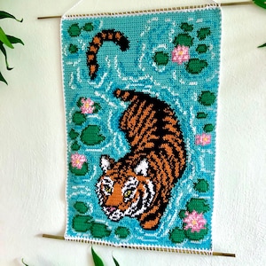 May include: A crocheted wall hanging featuring a tiger swimming in a blue pond with green lily pads and pink flowers.