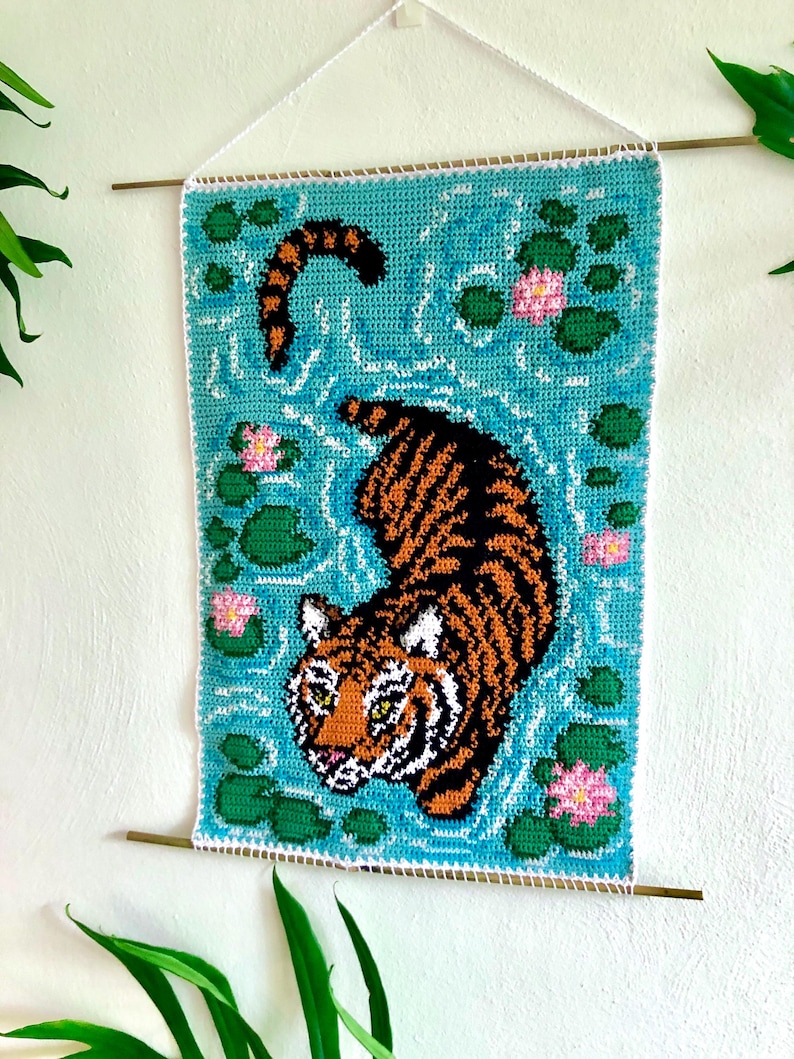 May include: A crocheted wall hanging featuring a tiger swimming in a blue pond with green lily pads and pink flowers.