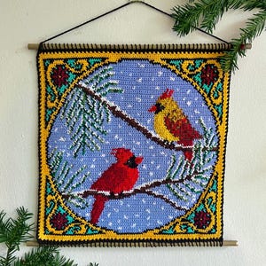 May include: A handmade wall hanging featuring two red and yellow cardinals perched on a snow-covered branch. The birds are set against a light blue background with white snowflakes. The border is yellow, black, and green, with a wooden dowel at the top.