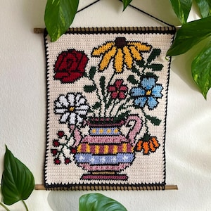 May include: A hand-crocheted wall hanging depicting a vase of flowers. The vase is pink with horizontal stripes. The flowers include a red rose, yellow daisies, and blue and white blooms. It has a black border and a wooden dowel for hanging.