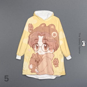 May include: Yellow hooded blanket with a cartoon bear design. The blanket features a large graphic of a character with glasses and a bear, with smaller bear illustrations. The cuffs and hood lining are white. The text "The Geeky Gamer" is visible.