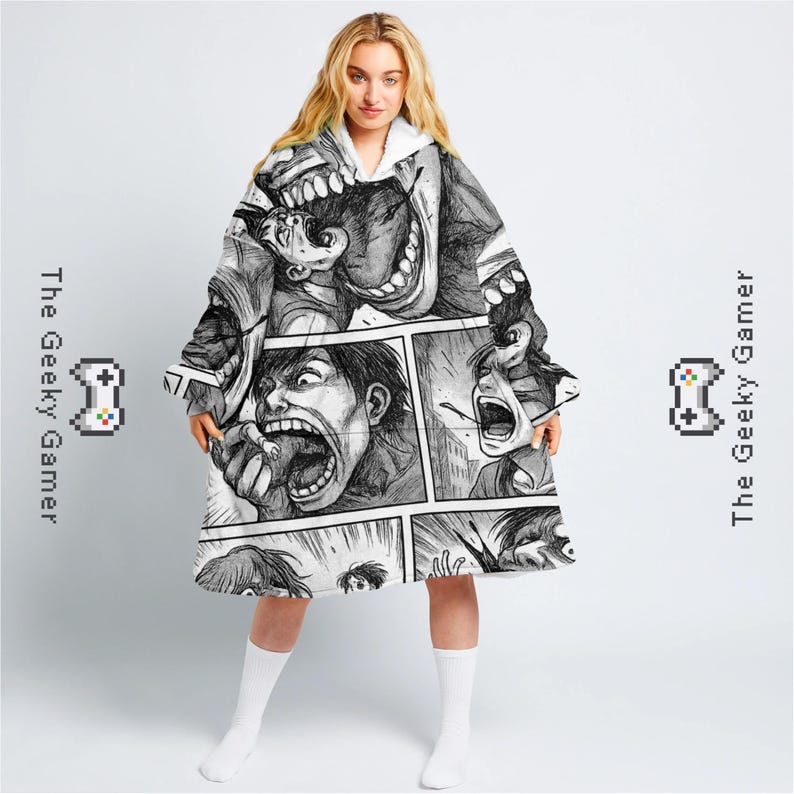 May include: A hooded blanket featuring a black and white manga-style comic print. The design showcases panels with screaming faces. The blanket has a white interior and is labeled with "The Geeky Gamer" and a pixelated controller graphic.