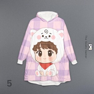 May include: A hooded blanket in a pink and purple plaid pattern, featuring a cartoon character with a bear-like hood. The character has brown hair, a red scarf, and a white bear hood with pink accents. The text "The Geeky Gamer" is on the right side.
