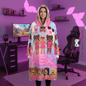 May include: A hooded blanket featuring a repeating pattern of cartoon characters in pink dresses and sunglasses. The blanket has a large front pocket and the name "ALEX" printed in pink. The background is a mix of light blue and pink.