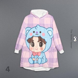 May include: A hooded blanket featuring a cartoon character in a blue bear costume. The blanket has a pink and purple plaid pattern and a white fleece-lined hood and cuffs. The text "The Geeky Gamer" is on the right side.