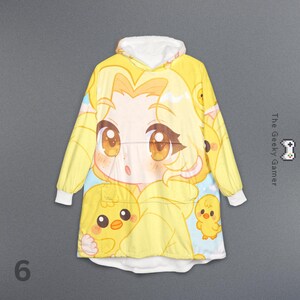 May include: Yellow hooded blanket featuring a large anime-style character with big brown eyes and yellow hair, surrounded by cartoon chicks. The blanket has a white fleece-lined hood and cuffs. The text "The Geeky Gamer" is on the right side.