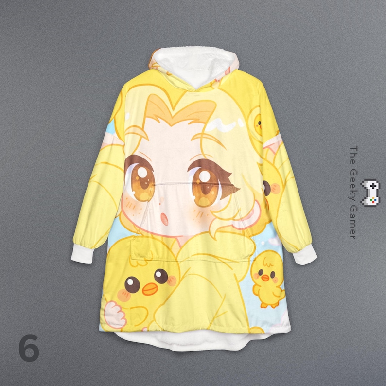 May include: Yellow hooded blanket featuring a large anime-style character with big brown eyes and yellow hair, surrounded by cartoon chicks. The blanket has a white fleece-lined hood and cuffs. The text "The Geeky Gamer" is on the right side.