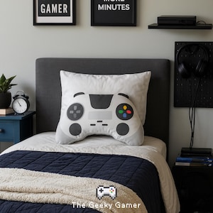 May include: A white pillow shaped like a video game controller rests on a bed with a navy blue comforter. The pillow has gray and black details, with colorful buttons. Above the bed are framed signs that read "GAMER" and "MORE MINUTES".