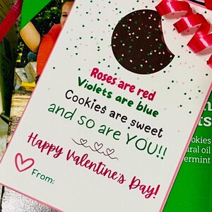 May include: A white card with pink and green hearts printed on it. The card has a cut-out shape of a chocolate chip cookie. The text on the card reads: "Roses are red, Violets are blue, Cookies are sweet, and so are YOU!! Happy Valentine's Day!"
