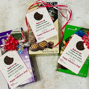 May include: Three Girl Scout cookie boxes with Valentine's Day gift tags attached. The tags are white with pink and red text that reads "Roses are red, Violets are blue, Cookies are sweet, and so are YOU!! Happy Valentine's Day!" Each tag has a chocolate cookie on it.