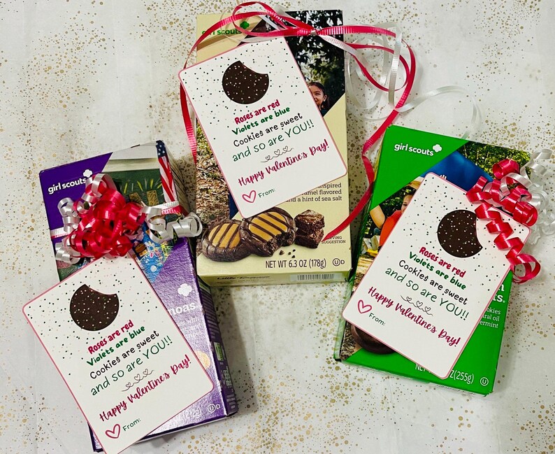 May include: Three Girl Scout cookie boxes with Valentine's Day gift tags attached. The tags are white with pink and red text that reads "Roses are red, Violets are blue, Cookies are sweet, and so are YOU!! Happy Valentine's Day!" Each tag has a chocolate cookie on it.