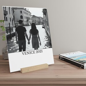 Può includere: Fotografia in bianco e nero di una coppia che cammina mano nella mano a Venezia, Italia, stampata su una carta bianca. La carta è sostenuta da un supporto in legno chiaro. Il testo "VENICE 2021" è stampato in basso.