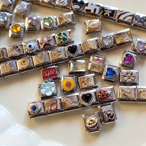 Puede incluir: Una colección de pulseras de abalorios plateadas y doradas con varios dijes decorativos. Los dijes incluyen corazones, flores, estrellas y letras. Algunos dijes tienen detalles coloridos.