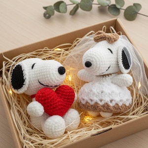 May include: Two handmade crochet Snoopy figures in a gift box. One Snoopy holds a red heart, the other wears a white dress and veil. The box is filled with straw and fairy lights, perfect for a wedding or anniversary gift.