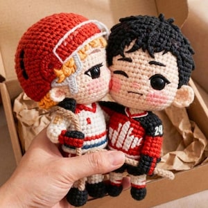 May include: Two crocheted dolls, one in a red hockey helmet and jersey, the other in a red hockey jersey with the number 24. The dolls are in a box, and the one on the right is winking. The dolls are holding hockey sticks.