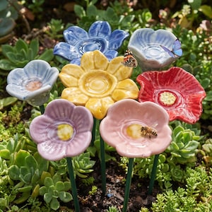 Könnte beinhalten: Eine Ansammlung von sieben blumenförmigen Gartensteckern aus Keramik in Blau, Gelb, Rot, Lavendel und Rosa, die jeweils auf einem dünnen grünen Metallstab befestigt sind. Ein Schmetterling sitzt auf der gelben, einer auf der hellblauen und eine Biene auf der rosa Blume.