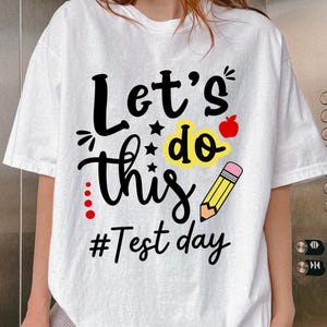 May include: White t-shirt with the phrase "Let's do this" in black script, accented with stars, a red apple, and a yellow pencil. The words "#Test day" are below the main text.