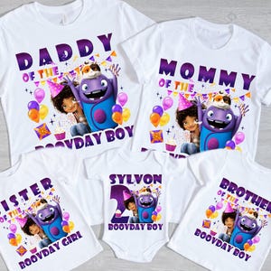 Puede incluir: Camisetas blancas y un body con gráficos coloridos. Las camisetas tienen texto que dice "Daddy", "Mommy", "Sister" y "Brother" con un personaje de dibujos animados y las palabras "Boovday Boy" o "Boovday Girl". Un body blanco dice "Sylvon 2 Boovday Boy".