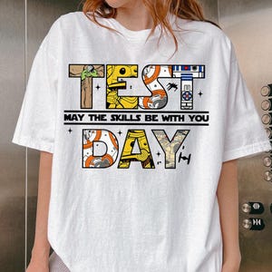 May include: White t-shirt with a Star Wars-themed graphic. The design features the words "TEST DAY" in a block letter style, with images of characters and symbols from the Star Wars franchise. The phrase "MAY THE SKILLS BE WITH YOU" is printed below.