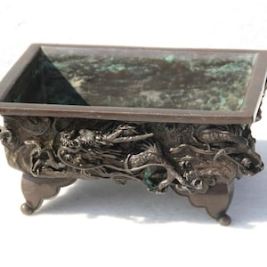 Può includere: Un vaso quadrato in bronzo con intricati disegni di draghi sui lati e sui piedi. Il vaso ha una patina verde ed è in buone condizioni.