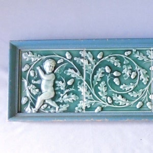 May include: A rectangular wall plaque featuring three green ceramic tiles set in a light blue wooden frame. The tiles display a raised white relief pattern of oak leaves, acorns, and a small cherub figure on the left side of the composition.