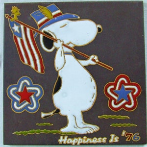 May include: A ceramic tile featuring Snoopy holding an American flag, wearing a patriotic hat, and surrounded by star-shaped designs. The tile has a brown background with the text "Happiness Is '76".
