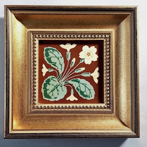 May include: A square decorative tile featuring a botanical illustration of a primrose plant with cream-colored flowers and green leaves on a dark red background, encased in a thick, ornate gold-toned frame with a beaded inner border detail.
