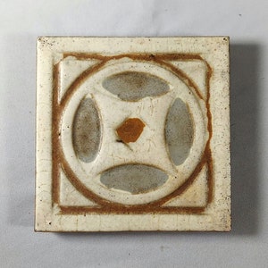May include: A square ceramic tile featuring a raised circular geometric pattern in the center. The tile has a cream-colored glaze with visible crackle textures and brown, rust-colored accents along the edges and within the central design.