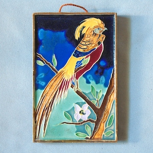 May include: Rectangular ceramic tile featuring a colorful bird perched on a branch. The bird has yellow, red, and blue plumage. The background is a gradient of blue and green. A small white flower is also visible.
