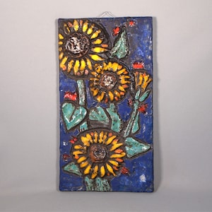 May include: A rectangular ceramic wall hanging featuring a sunflower design. The sunflowers have yellow petals and brown centers, set against a textured blue background. Green leaves and red accents complete the floral artwork.